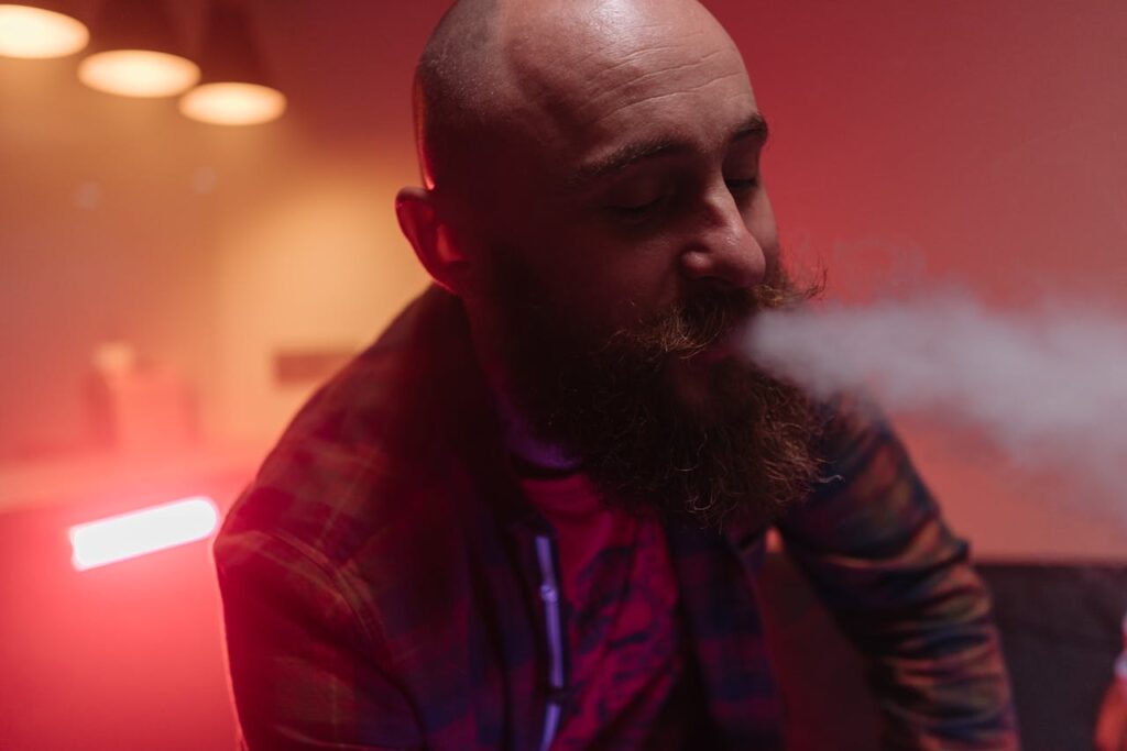 Close-up of a bald man with a beard exhaling vapor in a dimly lit indoor space.